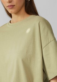 Lichtgroen oversized t-shirt met korte mouwen, voorzien van een kleine crèmekleurige zonnestraalafbeelding op de linkerborst. Zacht katoenen materiaal.