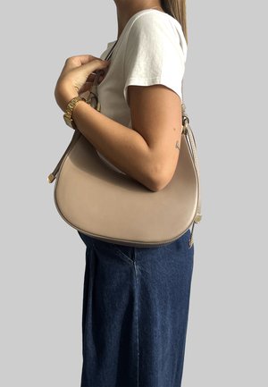 Borsa a mano in pelle beige con forma curvata, hardware dorato e una sola tracolla. Indossata con una gonna di jeans scura, esposta su uno sfondo grigio.