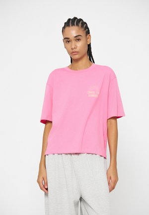 Jeune femme aux cheveux tressés portant un t-shirt rose oversize avec texte et un pantalon ample gris clair sur fond blanc.