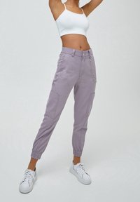 Pantalons cargo lilas avec des poignets élastiques, deux poches latérales et un détail en chaîne argentée. Associé à un crop top blanc et des baskets blanches.