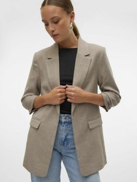 Beige blazer laget av teksturert stoff med en enkelt knapp foran, notched revers og to frontlommer. Båret over en svart topp og blå jeans.