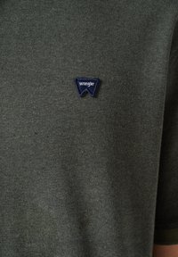 Tamsiai žalia marškinė su tekstūruotu audiniu, kurių kaklo priekyje yra mažas mėlynai juodas "Wrangler" logotipo užrašas. Trumpi rankovės.
