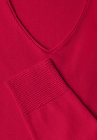 Haut tricoté rouge avec un design en V et des poignets à côtes, présentant une texture lisse avec des points réguliers.