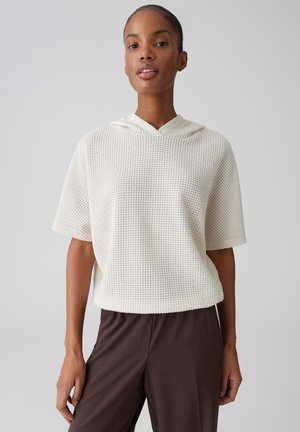 Frau trägt ein strukturiertes weißes, kurzärmeliges Kapuzenoberteil und locker sitzende dunkelbraune Hosen, steht vor einem einfarbigen hellgrauen Hintergrund.