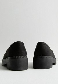 Mocasines de ante negro con una suela de goma negra gruesa, textura suave y un patrón en zigzag que acentúa el talón.