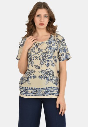 Donna con capelli castani ondulati che indossa una camicetta beige a maniche corte con motivi floreali blu e pantaloni larghi blu navy, in piedi davanti a uno sfondo bianco.