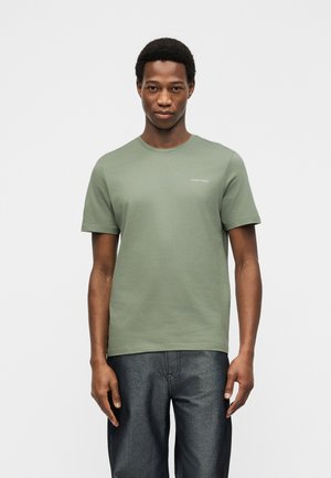 Noor mees seisab, vaadates otse ette, kandes helerohelist Calvin Kleini T-särki ja tumedat halli pükse, taust on ühevärviline.