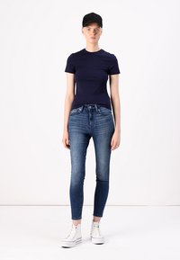Marineblå tilpasset t-skjorte kombinert med mørkeblå skinny jeans. Antrekket inkluderer hvite høyhælte joggesko og en svart caps. Enkelt design, minimale detaljer.