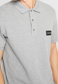 Polo en coton gris avec un petit col, trois boutons et un petit écusson noir affichant le nom de la marque sur la poitrine gauche.