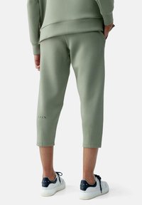 Pantaloni da jogging khaki a vita corta con una texture liscia e polsini elastici; presentano sottili ritagli laterali e una vestibilità aderente, abbinati a sneaker bianche.