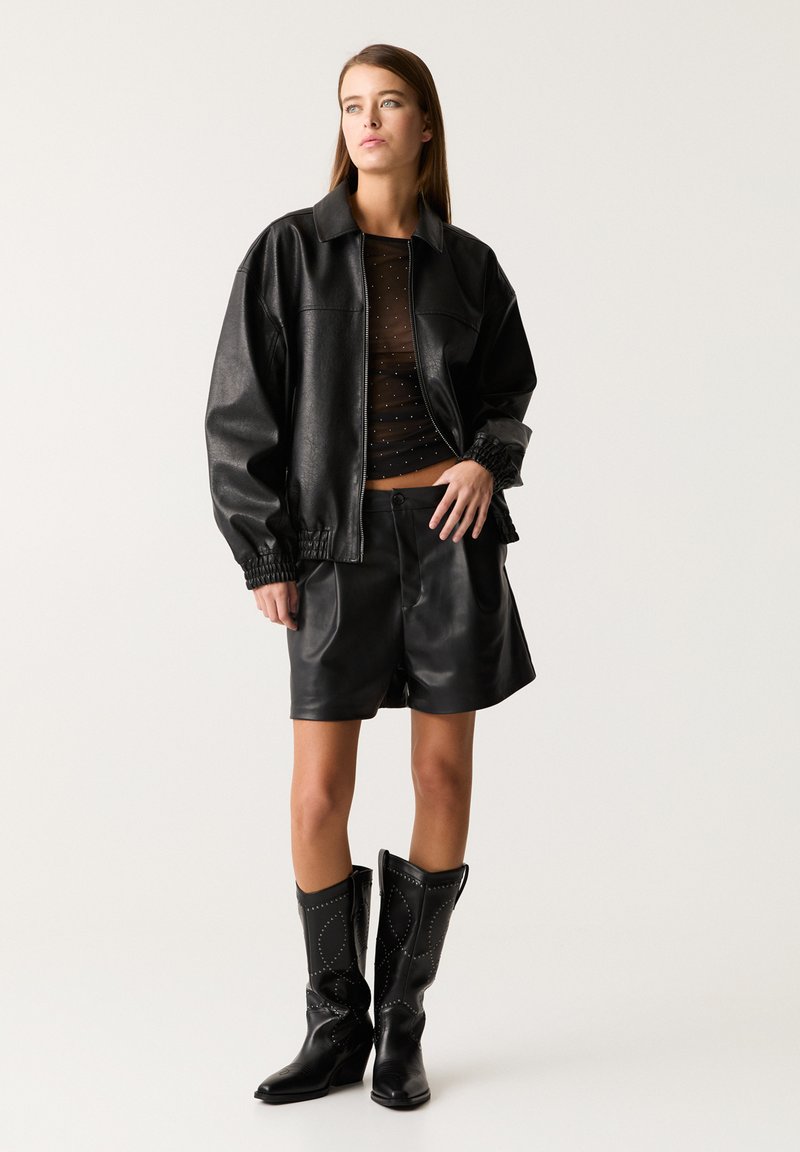 Donna che indossa un giubbotto in pelle sintetica nera, una maglia trasparente nera con pois, pantaloni corti in pelle sintetica nera e stivali neri con borchie fino al polpaccio.