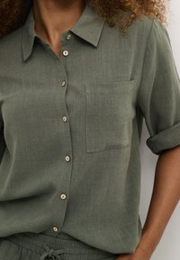Donna che indossa una camicia verde oliva con bottoni, munita di tasca sul petto e maniche arrotolate, infilata in pantaloni abbinati con vita regolabile tramite cordoncino.