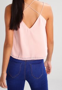 Ljusrosa ärmlös topp med V-formad rygg och korsande band, kombinerad med blå jeans som har bakfickor och sömdetaljer.