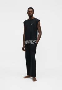 Vīrietis stāv basām kājām, ģērbies melnā bezroku topā ar baltu Moschino logotipu un melnās plašās biksēs uz baltā fona.