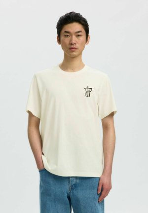 Jeune homme portant un t-shirt blanc uni à manches courtes avec un petit dessin de cafetière sur la poitrine gauche, associé à un jean bleu.