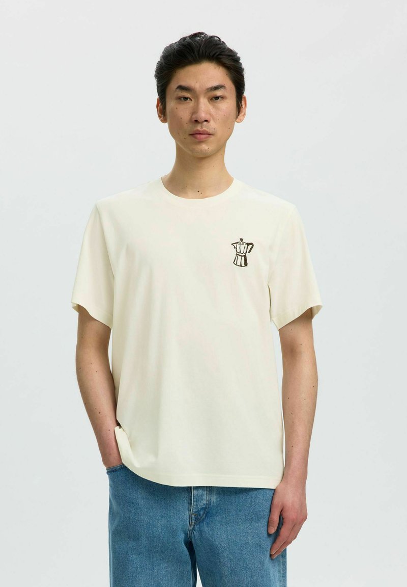 Jeune homme portant un t-shirt blanc uni à manches courtes avec un petit dessin de cafetière sur la poitrine gauche, associé à un jean bleu.
