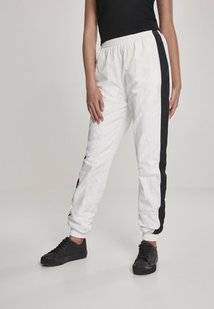 Hvite bukser med elastisk liv og svarte sidelister, strukturert stoff, med mansjetter ved anklene og en avslappet passform, vist sammen med svarte sneakers.