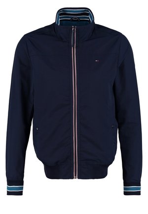 Homme posant en t-shirt bleu à motifs, veste zippée bleu marine, jean retroussé et baskets blanches sur un fond clair uni.
