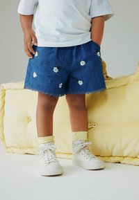 Denim shorts met witte madeliefjesborduursel, gegolfde zoom en zijzakken. Gecombineerd met witte hoge sneakers en gele sokken.