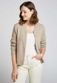 Cardigan en tricot beige avec texture côtelée, à manches longues et à ouverture frontale. Porté sur un haut blanc avec un pantalon crème ajusté.