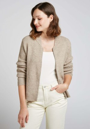 Cardigan en tricot beige avec texture côtelée, à manches longues et à ouverture frontale. Porté sur un haut blanc avec un pantalon crème ajusté.