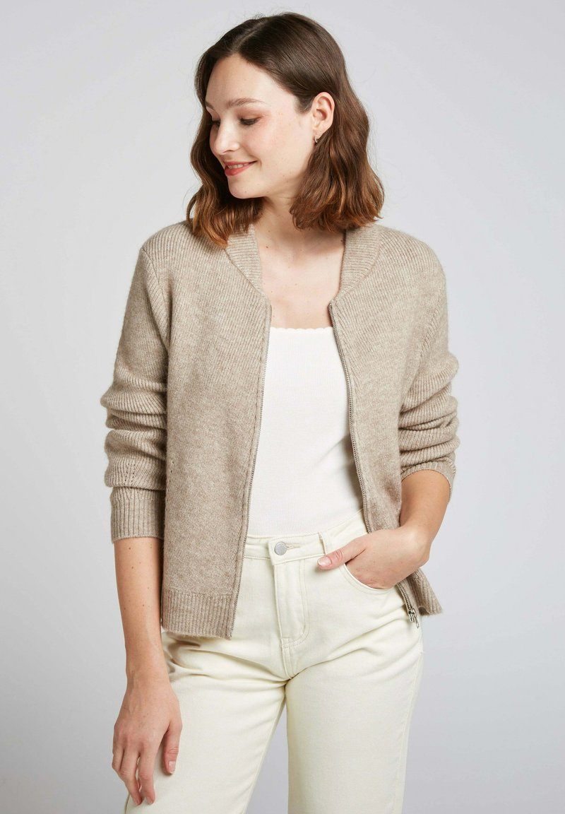 Cardigan en tricot beige avec texture côtelée, à manches longues et à ouverture frontale. Porté sur un haut blanc avec un pantalon crème ajusté.