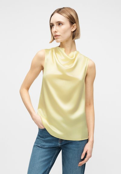 Γυναίκα με κοντά ξανθά μαλλιά που φοράει ένα αμάνικο ανοιχτόκκινο σατέν τοπ και μπλε τζιν, στέκεται με το ένα χέρι στην τσέπη.