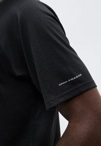 Κοντινό πλάνο του βραχίονα ενός ατόμου που φοράει μαύρο κοντομάνικο πουκάμισο με το λογότυπο "OMNI-FREEZE" τυπωμένο στο μανίκι.