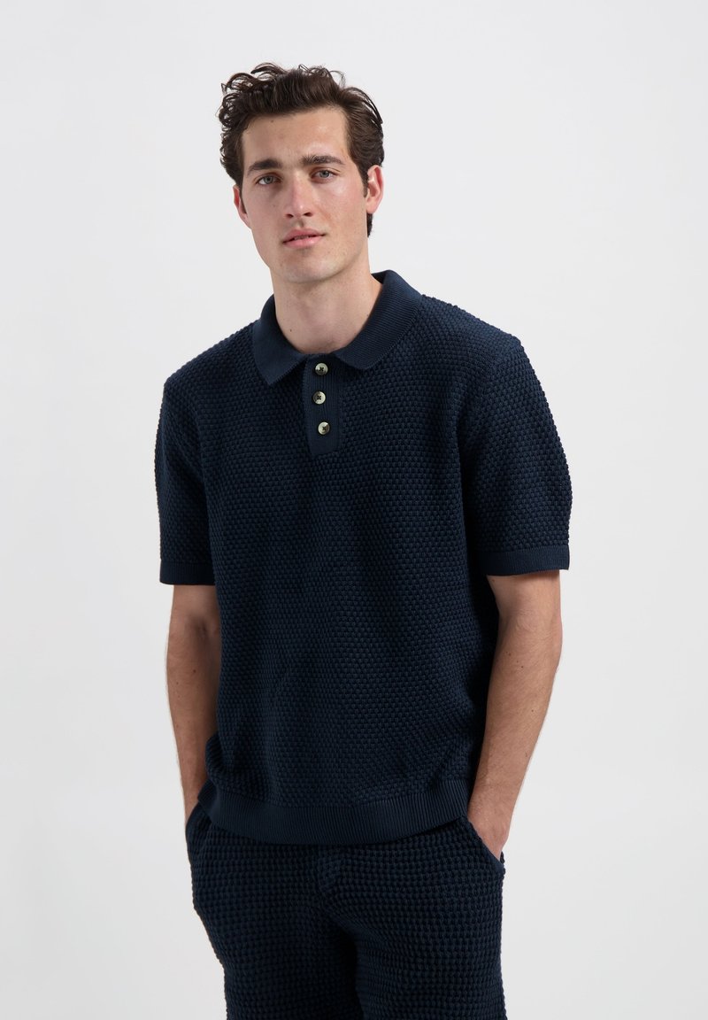 Marineblauw poloshirt met een gestructureerd, geweven patroon, voorzien van een geribbelde kraag en drie groene knopen bij de hals. Korte mouwen.