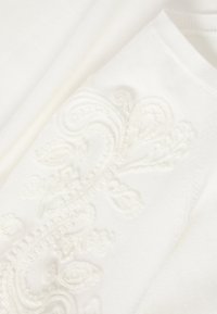 Λευκό ύφασμα με περίτεχνη ανάγλυφη κεντητική με λουλούδια και λεπτή υφή πλεκτού, που δείχνει λεπτομερείς ραφές και απαλές πτυχώσεις υλικού.