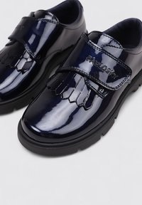 Chaussures en cuir verni bleu marine avec une bride Velcro, présentant un détail à franges sur le bout. Semelle épaisse en caoutchouc noir, offrant durabilité et soutien.