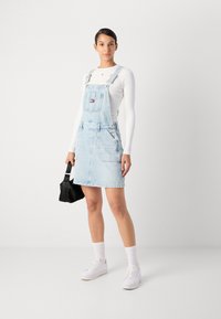 Denim jumpsuit i lys blå med justerbare stropper, frontlomme og detalj på sidelommen. Båret over en hvit langermet skjorte.