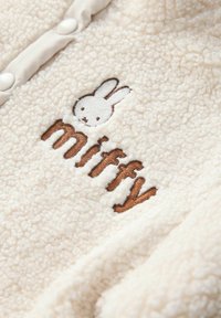Tissu en polaire crème doux avec le texte "miffy" brodé en marron et un petit visage de lapin blanc au-dessus.
