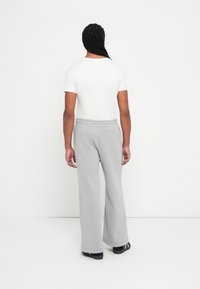 Person med langt flet hår iført en hvid t-shirt, grå joggebukser og sorte slip-on sko, stående foran en hvid væg.