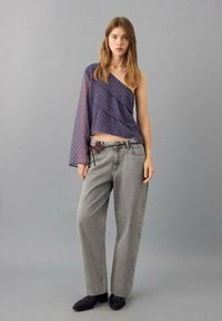 Fialový vzorovaný top s jedním odhaleným ramenem a dlouhým rukávem, kombinovaný se šedými širokými džíny a černými kotníkovými botami. Jednoduchý opasek s květinovým vzorem.