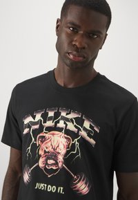 Sort bomulds t-shirt med et iøjnefaldende grafik af en bulldog med en vægtstang og lynakcenter, med teksten "NIKE" og "JUST DO IT".