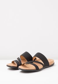 Svarta sandaler med två breda tygremmar och en smal rem, med en formad brun fotbädd och en flexibel svart sula.