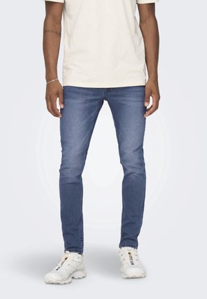 Mann trägt slim-fit blaue Jeans, weiße Sneakers und ein cremefarbenes T-Shirt und steht vor einem schlichten Hintergrund.