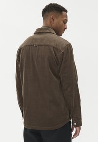 Pruunist kordurooist särgijope, millel on krae, vertikaalne ribakujundus ja metallist aktsent seljal. Nööpidega varrukad.