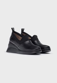 Zapatos negros sin cordones con parte superior de cuero texturizado, punta redonda y suela en cuña. El diseño presenta un suave acento de tela en la abertura.