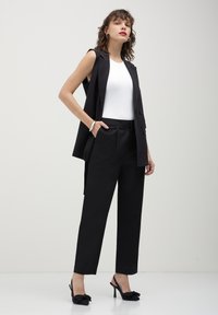 Femme debout en haut sans manches blanc, blazer sans manches noir, pantalon noir, talons noirs avec nœuds, et bracelet en argent au poignet.