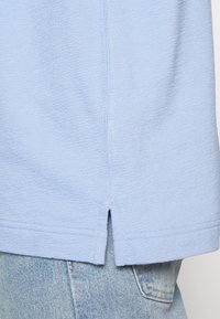 Lichtblauw katoenen t-shirt met een gestructureerde stof, met een subtiele split aan de onderkant en gestikte details. Draagt over lichte denim.