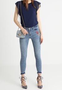 Top azul marino con mangas acampanadas, vaqueros de corte ligero con bordados y zapatos de tacón de diseño. Pequeño bolso cruzado gris.