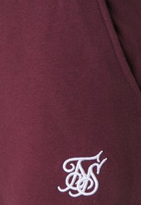 Burgundy sweatshirt tillverkad av mjukt tyg, med en vit broderad logotyp på den nedre vänstra sidan. Slät yta med en avslappnad design.