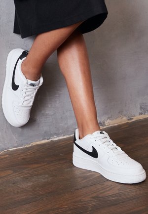 Nogi skrzyżowane w kostkach, w białych butach sportowych Nike z czarnymi logo, stojące na drewnianej podłodze przy teksturowanej szarej ścianie.