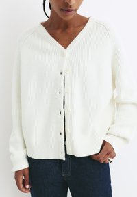 Weißer gerippter Cardigan mit V-Ausschnitt, Knopfverschluss und langen Ärmeln. Texturiertes Material und lockere Passform. Über dunklen Jeans getragen.