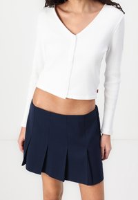Cardigan corto a maniche lunghe con costine bianche e chiusura a bottoni, abbinato a una mini gonna plissettata blu navy. Tessuto morbido e vestibilità su misura.