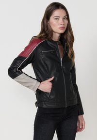 Giubbotto di pelle nera con accenti rossi e grigi, dotato di un colletto alto, tasche con zip e texture a coste sulle spalle. Silhouette slim fit.