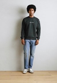 Donkergrijze sweatshirt met een ronde hals en witte tekst, gecombineerd met lichtblauwe jeans en grijze sneakers met groene accenten.