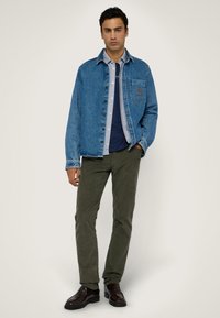 Jasnoniebieska kurtka dżinsowa z zapięciem na guziki z przodu oraz kieszenią na piersi; zestawiona z oliwkowo-zielonymi spodniami i ciemnymi butami eleganckimi.